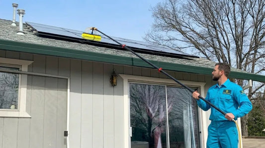 Professional cleaner in a blue uniform uses a long carbon water fed pole with a brush head to clean solar panels along a residential roofline while standing safely on the ground.