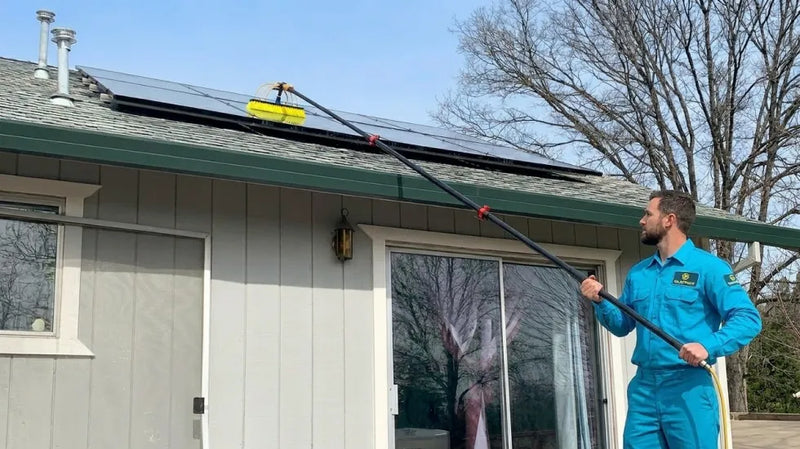 Professional cleaner in a blue uniform uses a long carbon water fed pole with a brush head to clean solar panels along a residential roofline while standing safely on the ground.