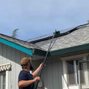 A man using the extended water fed pole to clean a solar panel on a roof, demonstrating its reach and effectiveness.